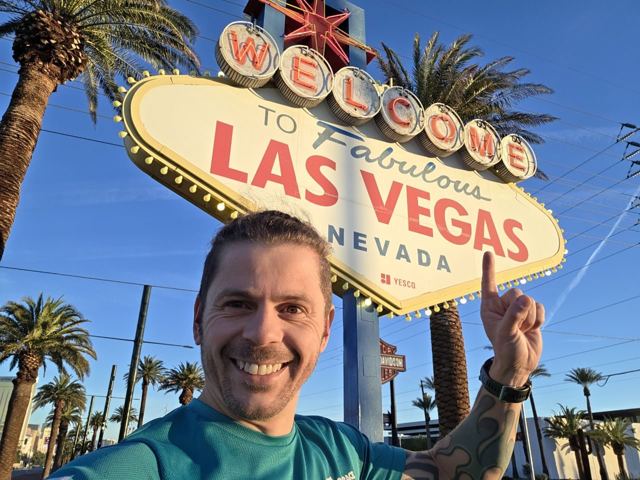 A man pointing at the famous Welcome to Fabulous Las Vegas sign under a blue sky.