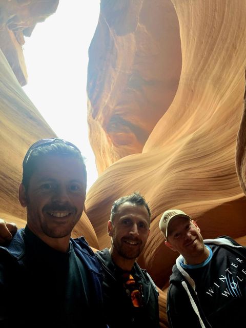 Three men taking a selfie among glowing waves of orange sandstone in a slot canyon.