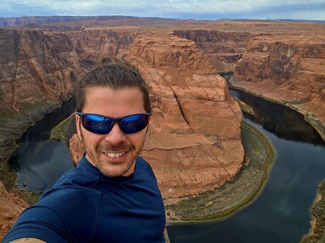 A man in sunglasses taking a selfie above the huge curve of Horseshoe Bend.
