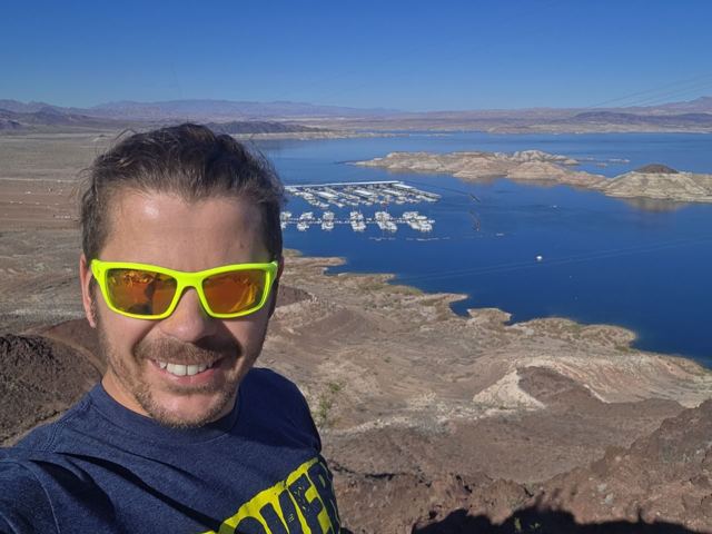 A man in yellow sunglasses taking a selfie above a blue lake in the desert.
