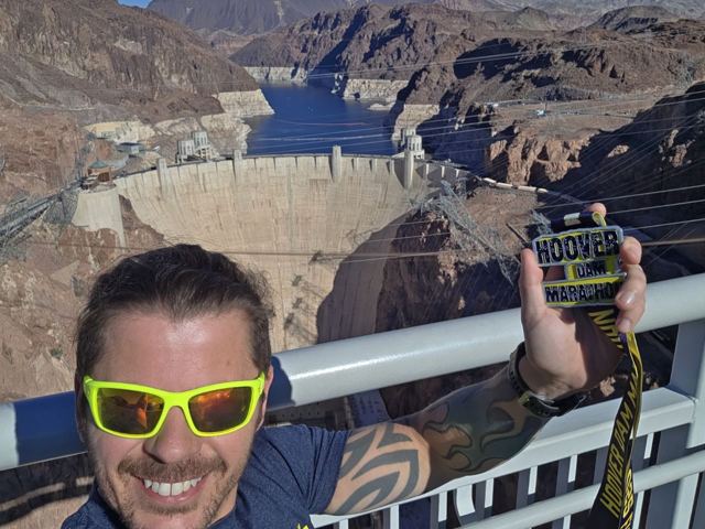 A man in yellow sunglasses holding a race medal with the Hoover Dam behind.