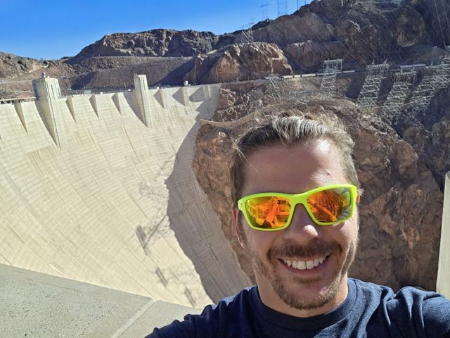 Smiling man in orange-lensed sunglasses taking a selfie in front of the Hoover Dam's concrete face.