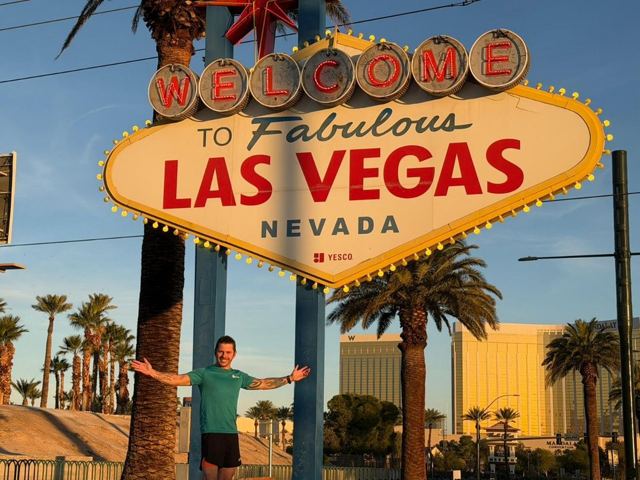 Man posing with arms outstretched beneath the Welcome to Fabulous Las Vegas sign, palm trees around him.