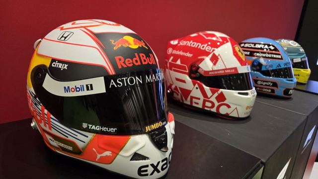 Row of colourful Formula 1 racing helmets displayed on a shelf against a red wall.