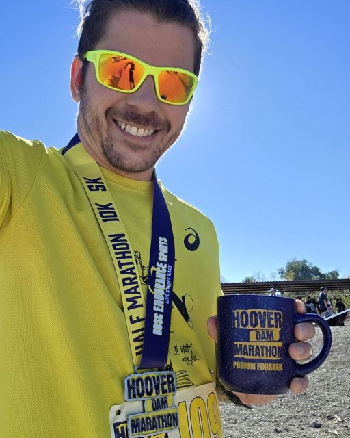 Smiling man in a yellow top and sunglasses holding a Hoover Dam Marathon mug and wearing a half marathon medal.
