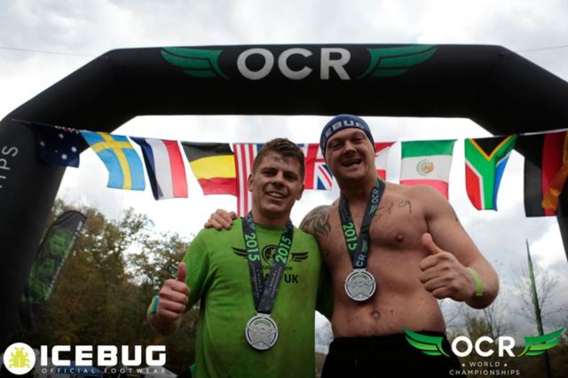 Two men in race tops showing their medals under the OCR World Championships finish arch.