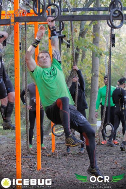 A man in a green top swinging across hanging rings on an obstacle course in a forest.