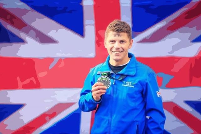 A man in a blue Team GB jacket holding a medal in front of a large Union flag.