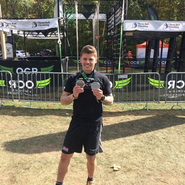 A man in a black race top holding up two medals in the OCR World Championships race village.
