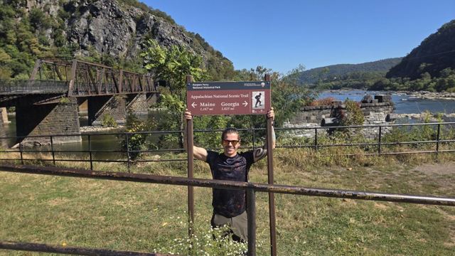 A man standing by a trail sign above a river with wooded hills and a bridge.