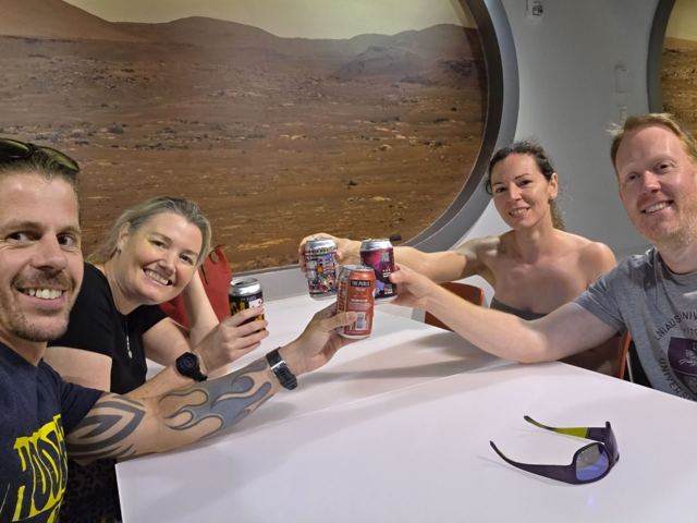 Friends raising cans of drink at a table in front of a Mars landscape mural.