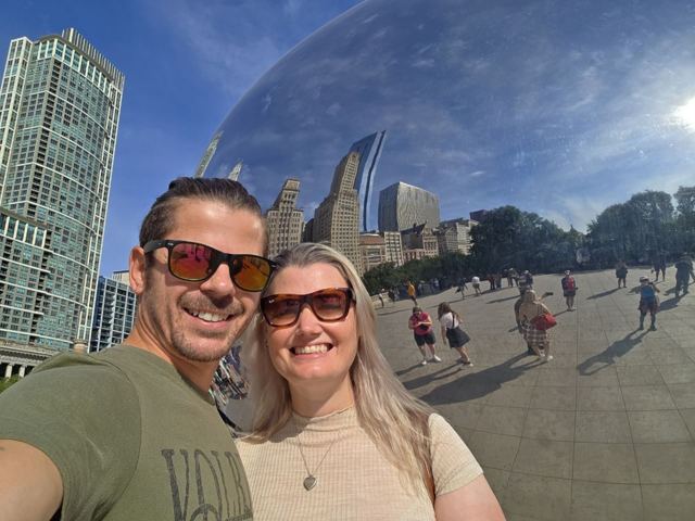 A man and a woman in sunglasses taking a selfie reflected in Cloud Gate.