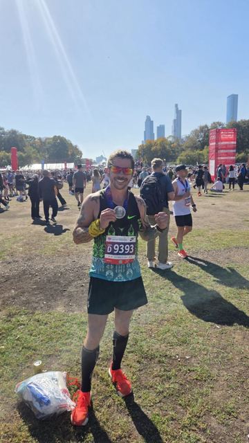 A runner with a race number holding up his medal in a busy park after the race.