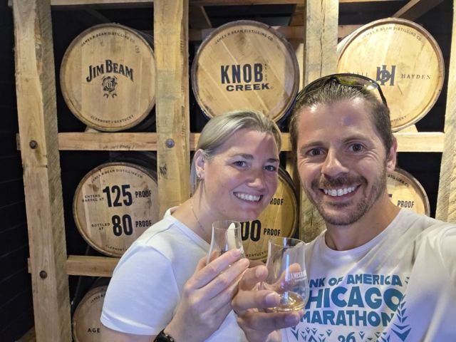 A man and a woman smiling with tasting glasses in front of stacked bourbon barrels.