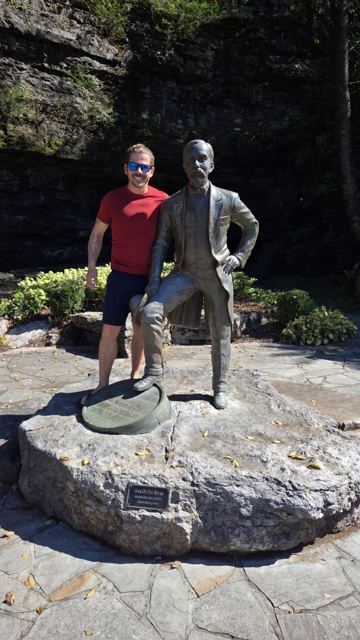 A man standing beside a bronze statue of Jack Daniel in front of a rocky cave.