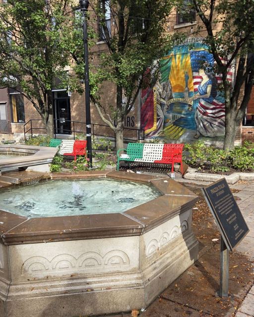 A stone fountain in a small leafy square with a bench painted in Italian flag colours and a mural.