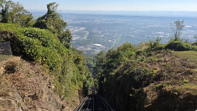 Steep view down a wooded mountainside to a wide city and plain far below.