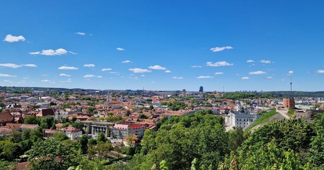 Panoramic view over Vilnius with red rooftops, spires and green trees under a blue sky.