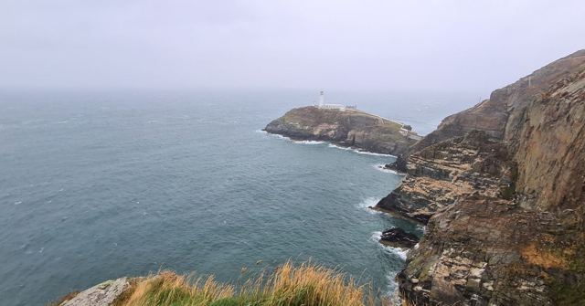 A white lighthouse on a rocky islet below steep sea cliffs, with calm grey sea and sky.