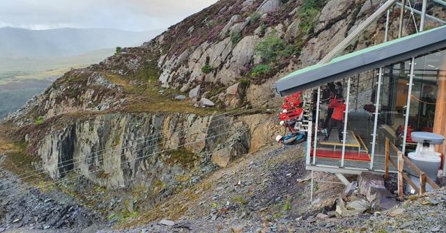 A small glass-fronted building on the edge of a steep grey slate quarry with hills behind.