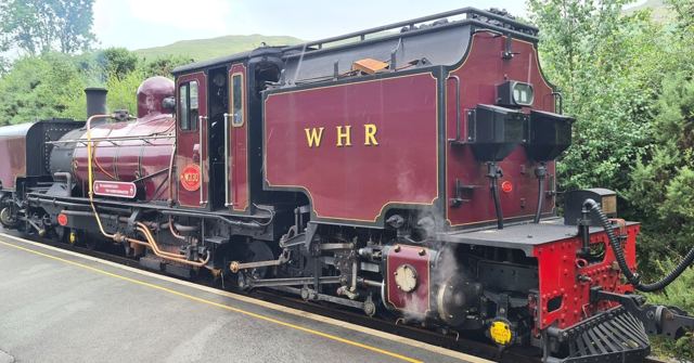 A maroon Welsh Highland Railway steam locomotive standing beside a platform with trees behind.