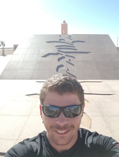 A man in sunglasses taking a selfie in front of a monument with a plane silhouette.