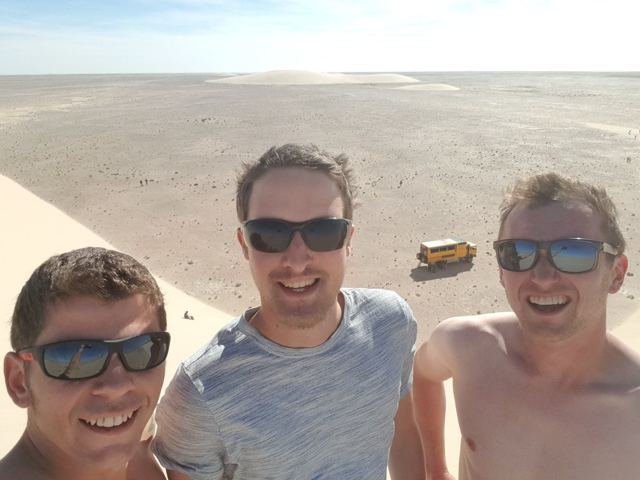 Three men in sunglasses taking a selfie on top of a sand dune above a desert plain.