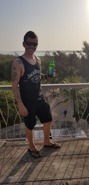 A man in a vest holding a bottle on a balcony overlooking a river.