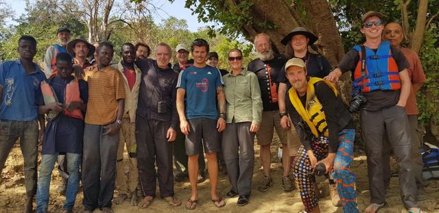 A large group of travellers and local people standing together under trees.