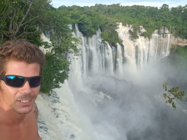 A man in sunglasses taking a selfie in front of the wide, misty Kalandula Falls.