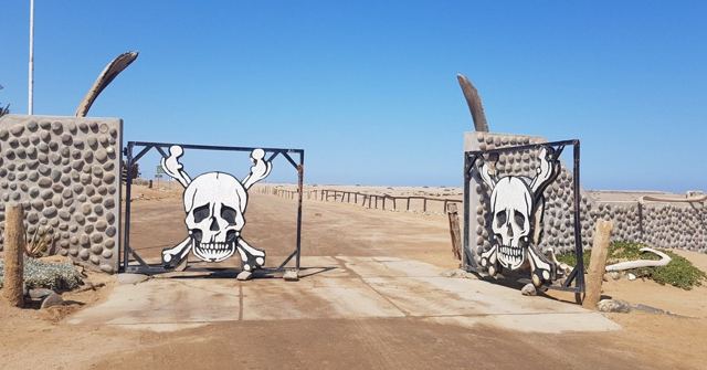 A park entrance gate decorated with large skull and crossbones on a sandy road.