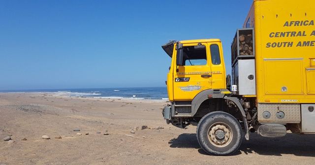 A yellow overland truck parked on a sandy beach next to the ocean.