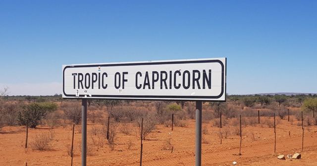A white Tropic of Capricorn road sign on red earth with scrub behind.