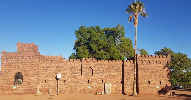 An old red sandstone building with battlements next to a palm tree and green tree.