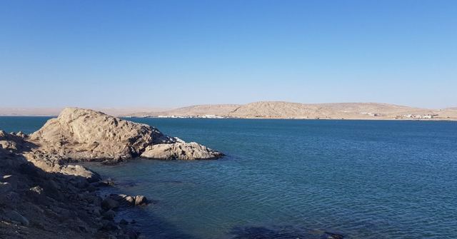 A rocky outcrop in a deep blue bay with sand dunes on the far shore.