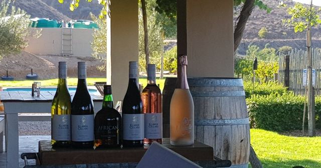 Several wine bottles lined up on a counter next to a barrel with a garden behind.