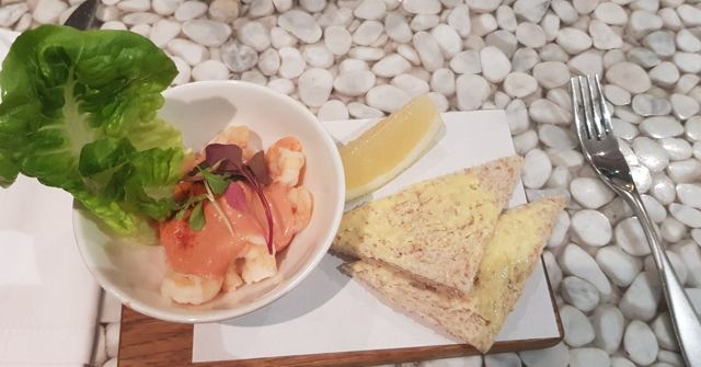 A bowl of prawn salad with lemon and a slice of toast on a pebble-patterned table.