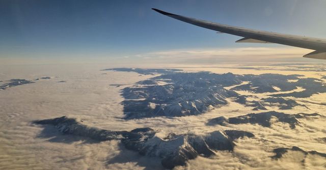 Snowy mountain ridges rising above a layer of cloud, seen from a plane window with the wing visible.