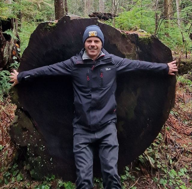 A man in a beanie standing arms outstretched in front of a giant cut tree trunk in a forest.