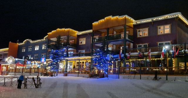 Colourful ski village buildings decorated with festive lights at night above a snowy street.