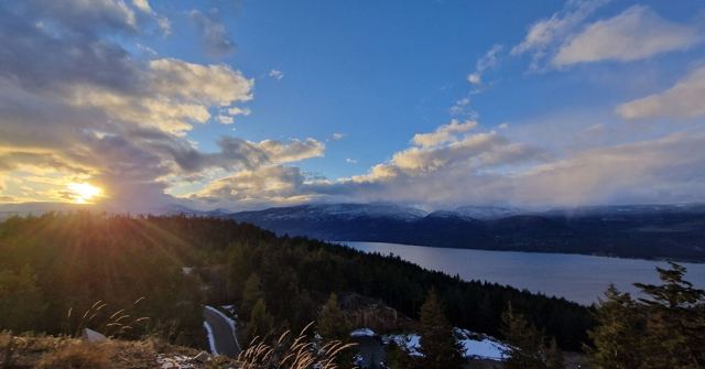 The sun setting behind forested hills above a lake, with dramatic clouds lit gold.