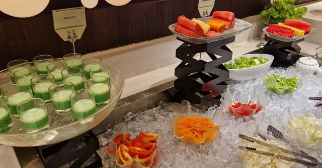 A hotel breakfast buffet with sliced watermelon, papaya, salad and rows of green jelly cups on ice.