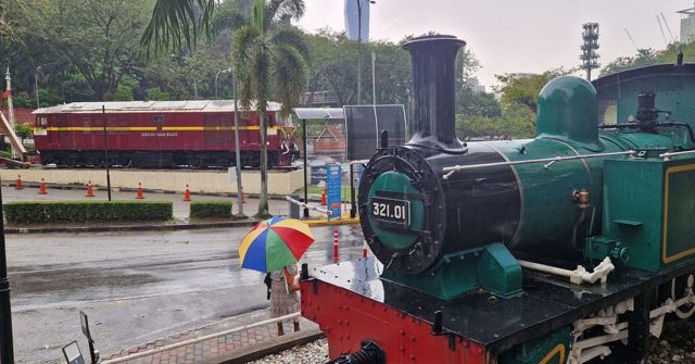 A vintage green steam locomotive displayed beside a red diesel train on a rainy day in Kuala Lumpur.