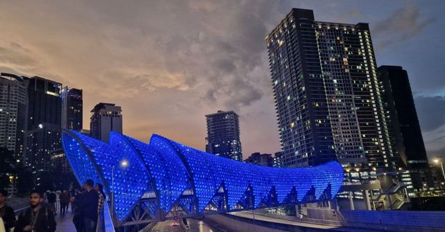 A curved pedestrian bridge lit bright blue at dusk with Kuala Lumpur skyscrapers behind.