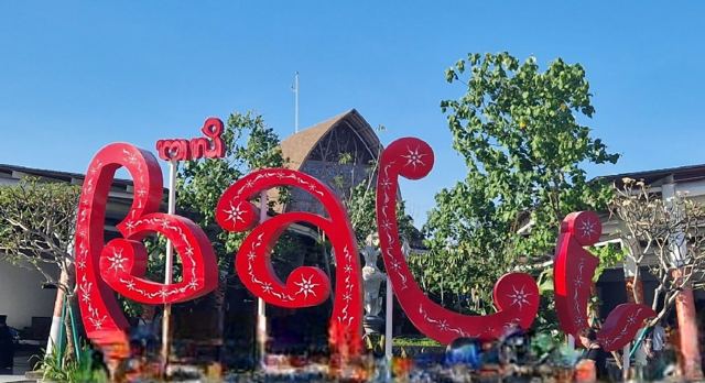 Large red curly letters spelling Bali in front of a traditional-style building and trees.