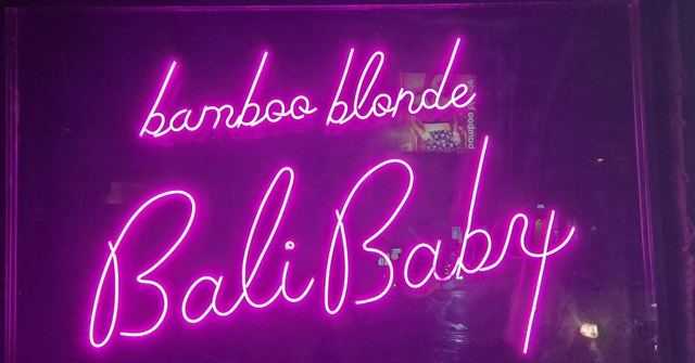 A pink neon sign reading bamboo blonde Bali Baby on a dark purple wall.