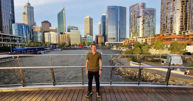 A man standing on a waterfront walkway with Perth's glass skyscrapers behind him.