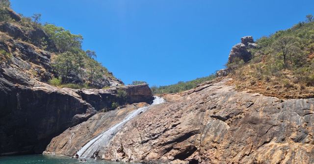 A waterfall sliding down a sloping granite rock face into a pool under a blue sky.