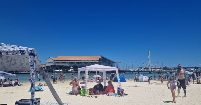 A busy sandy beach with a gazebo and beachgoers beside a harbour under deep blue sky.