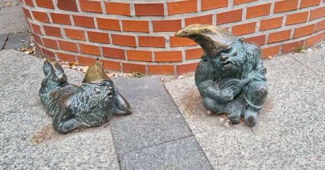 Two small bronze dwarf statues lounging on the pavement beside a brick wall.